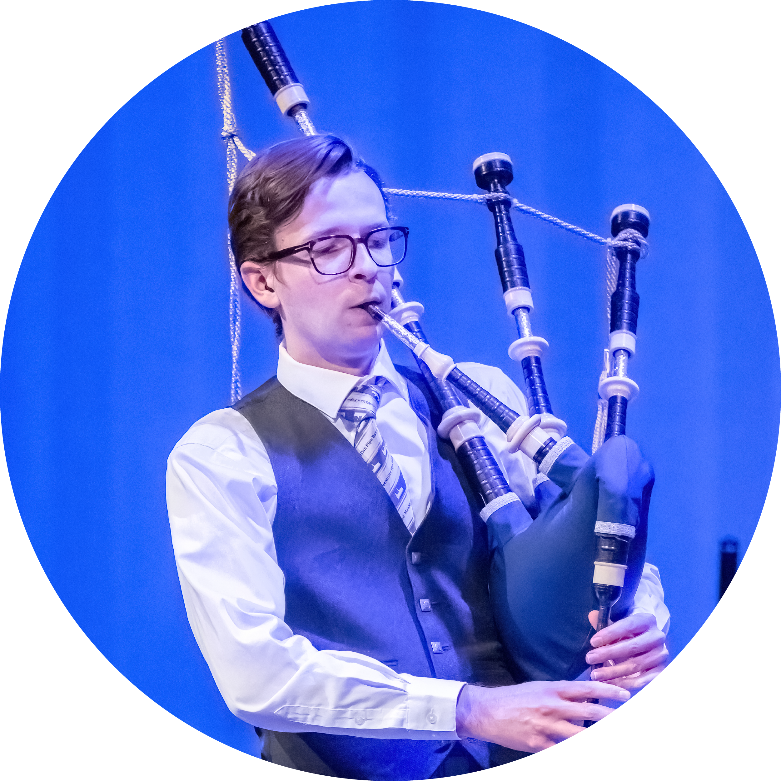 A man with short hair and glasses wearing a black vest with a dress shirt and tie. He is playing the bagpipes.